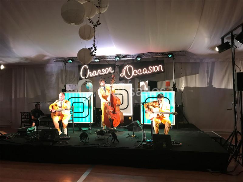 Musiciens jouant sur scène sous une tente éclairée, avec un panneau lumineux ’Chanson d'Occasion’ en arrière-plan.