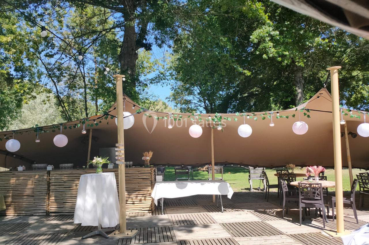 Espace extérieur aménagé avec tables, chaises et guirlandes lumineuses sous une tente beige, entouré de verdure.