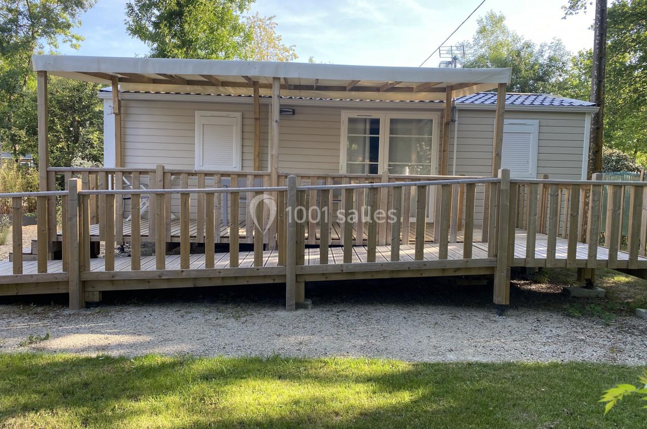 Mobil-home beige avec terrasse en bois couverte, entouré d'arbres et d'une pelouse.