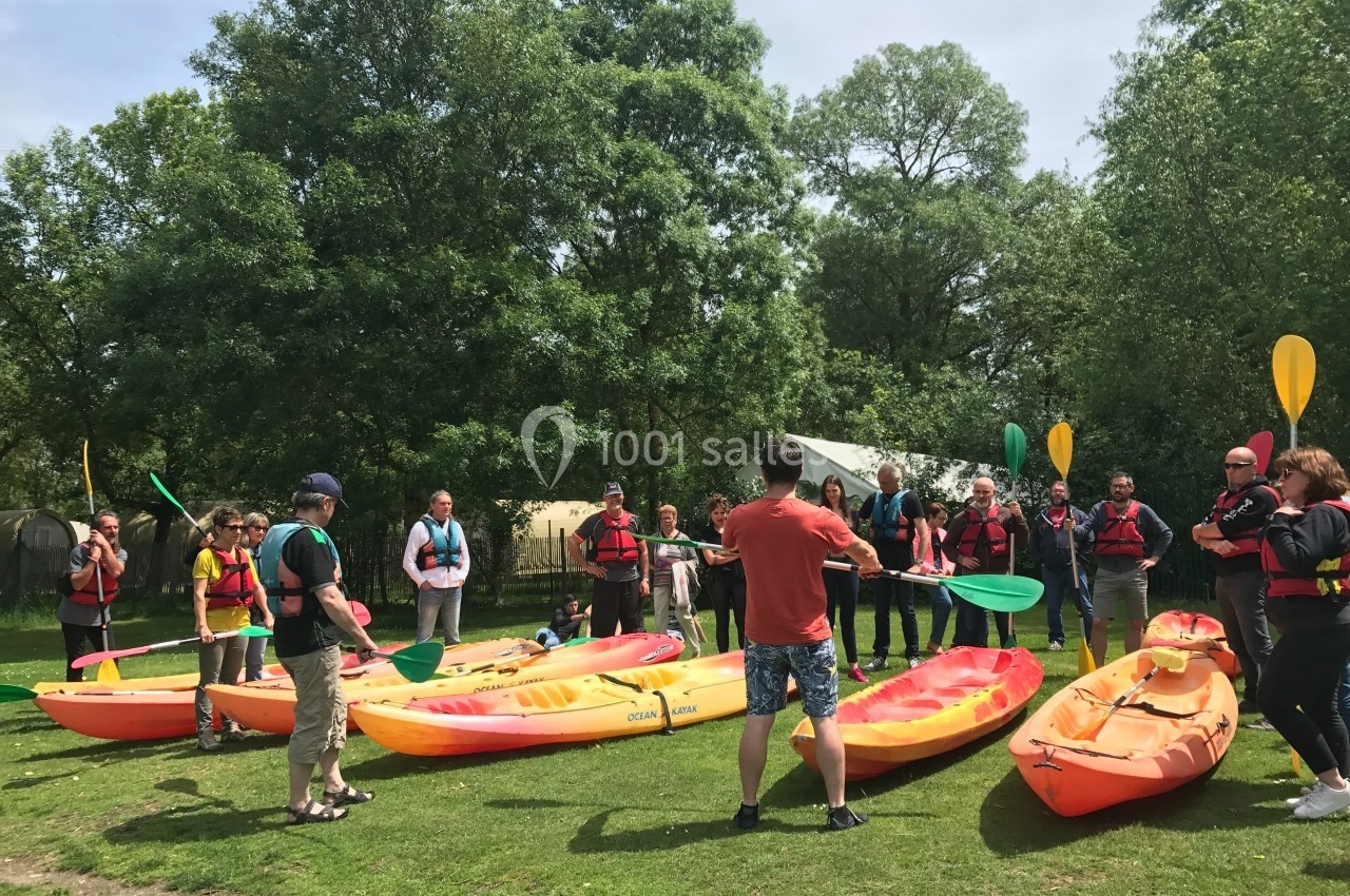 Un groupe de personnes en gilets de sauvetage écoute des instructions près de kayaks sur une pelouse verdoyante.