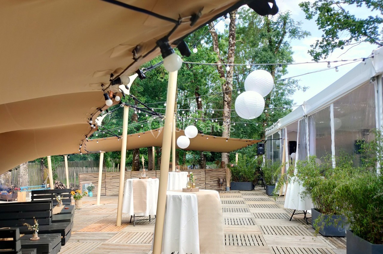 Terrasse extérieure avec tente beige, lanternes suspendues et tables hautes, entourée de végétation et d'une structure…