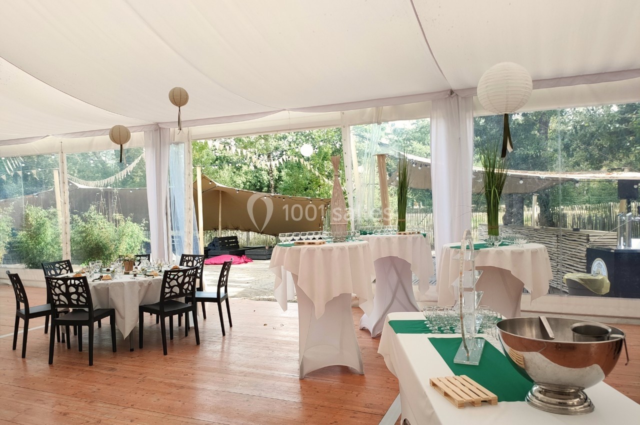 Salle de réception sous une tente avec tables dressées, nappes blanches et vue sur un espace extérieur arboré.