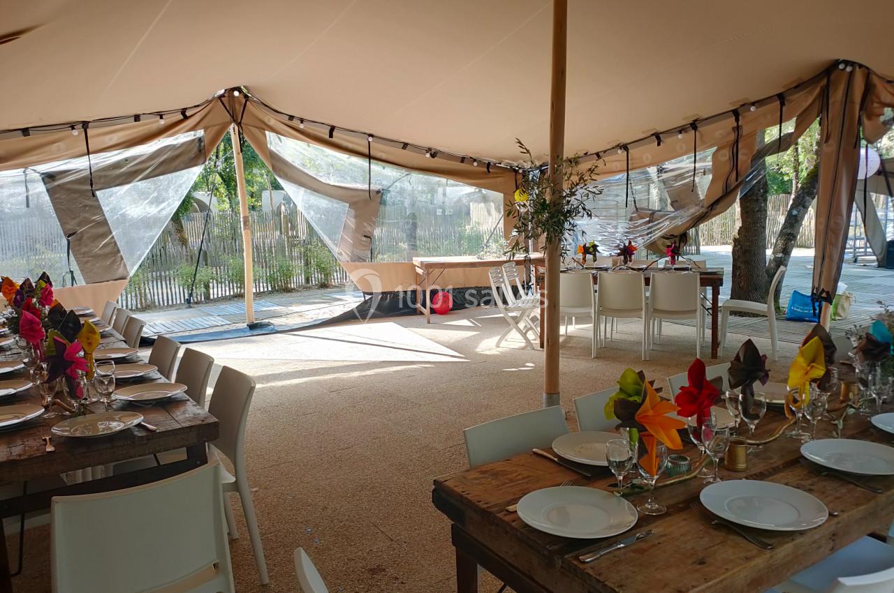 Intérieur d'une tente aménagée avec des tables en bois dressées pour un repas, décorées de serviettes colorées.
