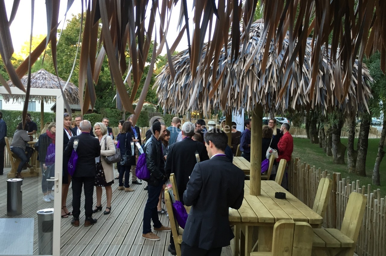 Groupe de personnes discutant sur une terrasse en bois, entourée de parasols en feuilles et d'arbres.