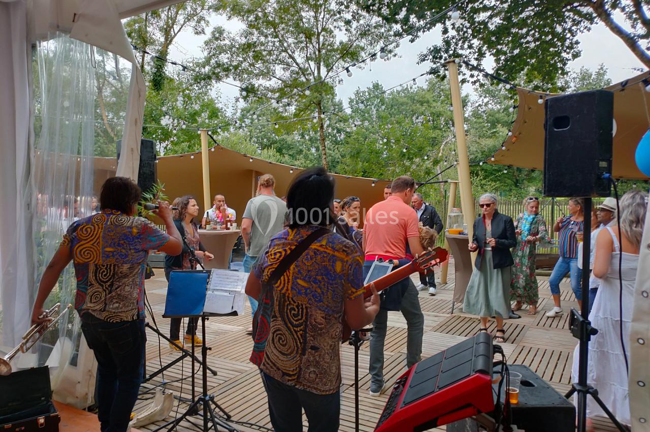 Groupe de musiciens jouant en plein air devant un public dans un espace boisé avec des décorations suspendues.