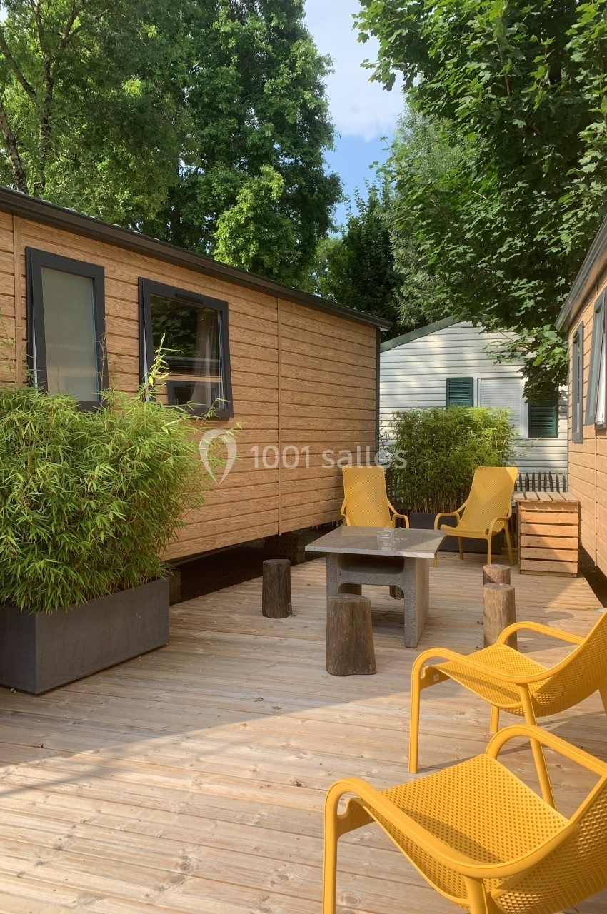 Terrasse en bois avec table basse, chaises jaunes, bambou en pot et mobil-homes en arrière-plan sous des arbres.