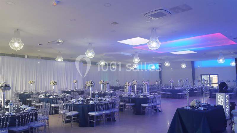 Salle de réception élégante avec tables dressées, chaises transparentes et éclairage moderne aux tons bleus et blancs.