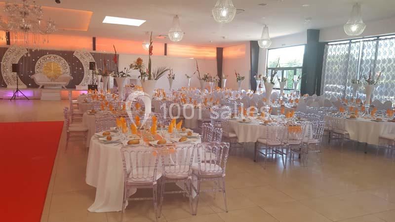 Salle de réception décorée avec des tables rondes, nappes blanches, chaises transparentes et serviettes orange.