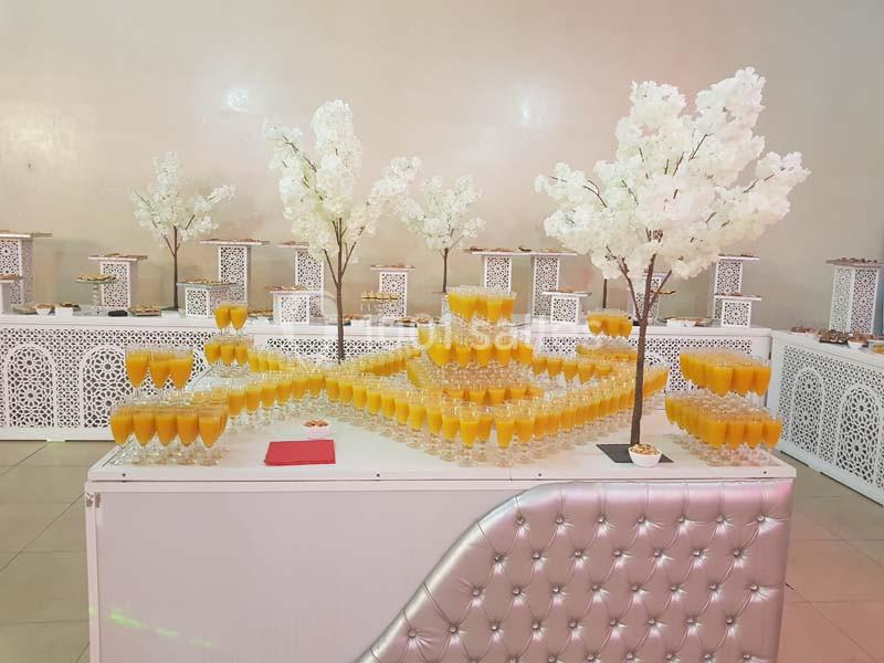Buffet décoré avec des arbres artificiels blancs et des verres de jus orange disposés en pyramides sur une table blanche.