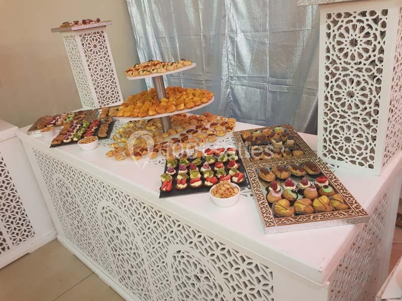 Table garnie de pâtisseries variées, présentées sur des plateaux et un présentoir, dans un décor blanc ajouré.