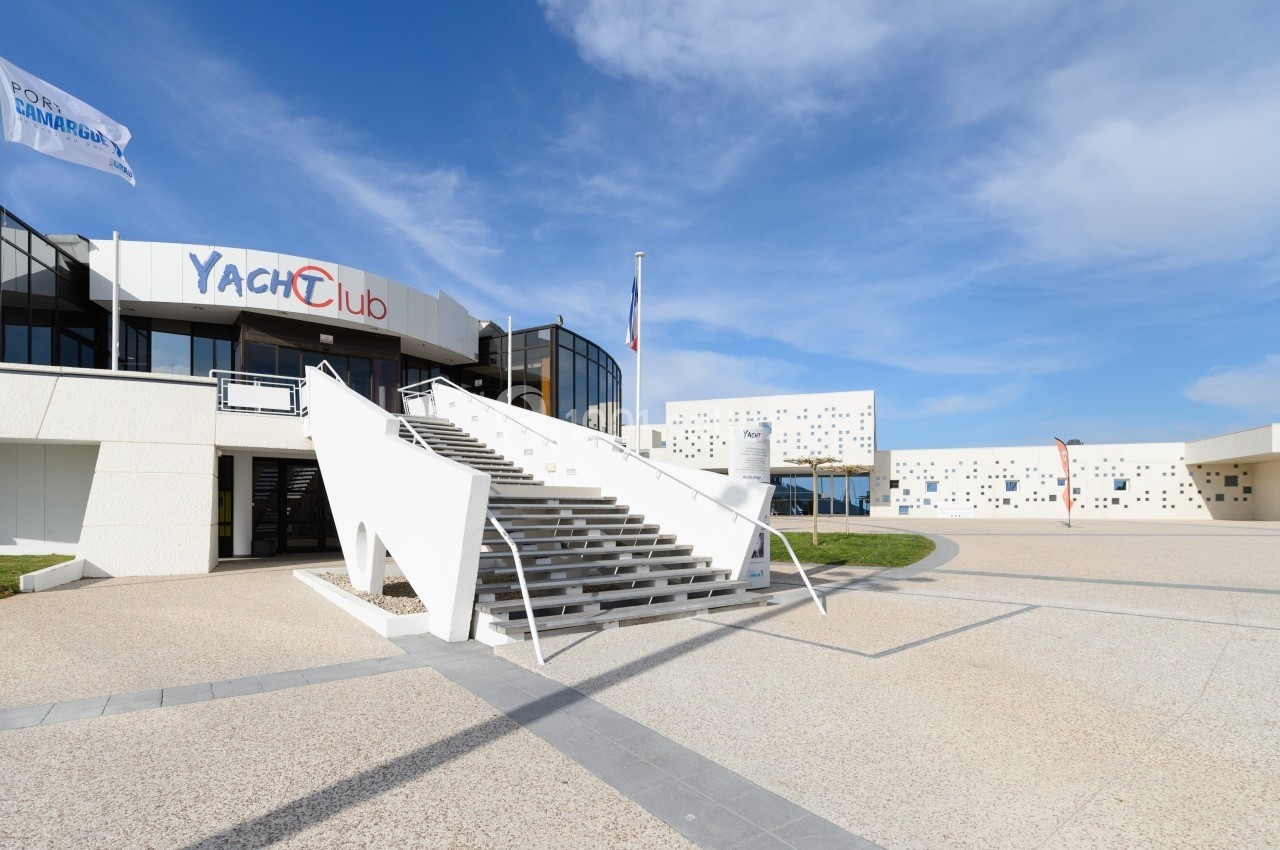 Escalier menant à l'entrée d'un bâtiment moderne marqué ’Yacht Club’, avec une place pavée et un ciel dégagé.