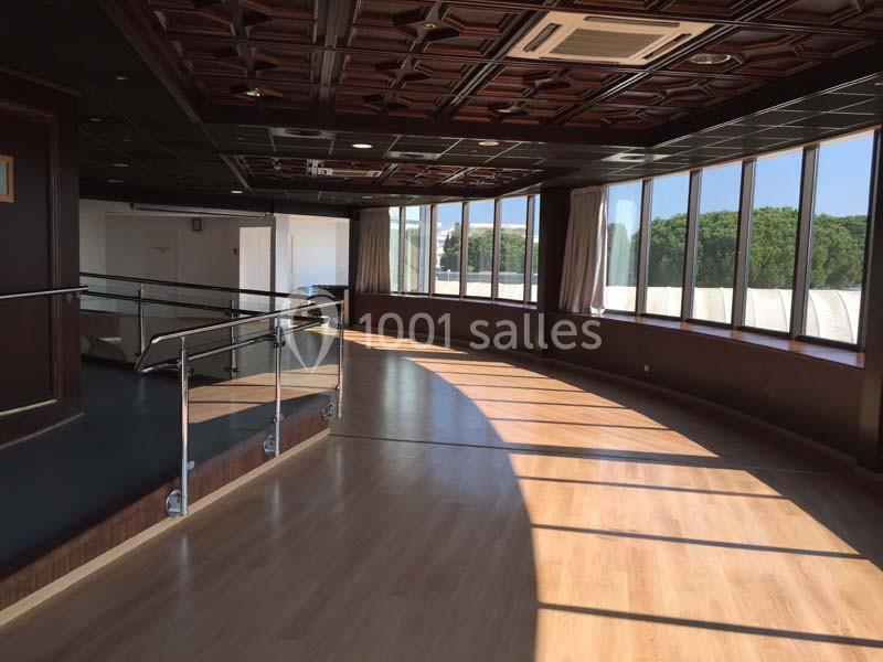 Salle lumineuse avec parquet, grandes baies vitrées courbées, plafond en bois sculpté et rampe métallique.