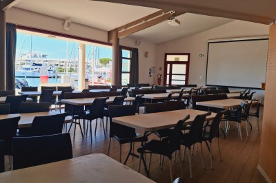 Terrasse d'un restaurant en bois avec vue sur un port de plaisance, tables occupées et végétation en premier plan.