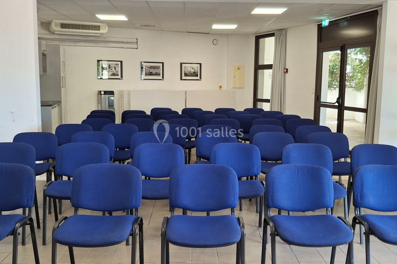 Salle de réunion avec des rangées de chaises bleues vides, éclairée par la lumière naturelle provenant des fenêtres.