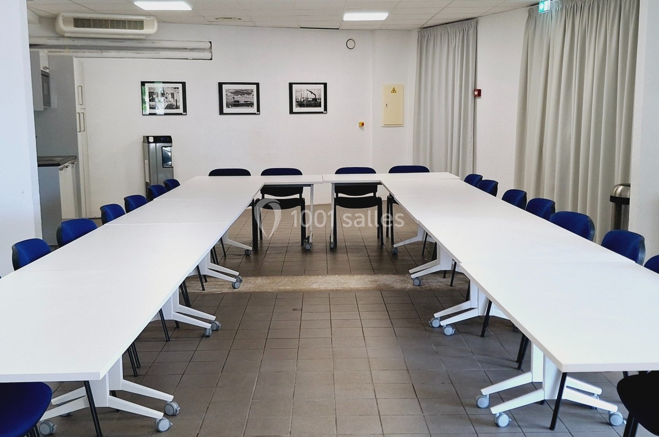 Salle de réunion lumineuse avec tables blanches disposées en U, chaises bleues et cadres accrochés au mur.