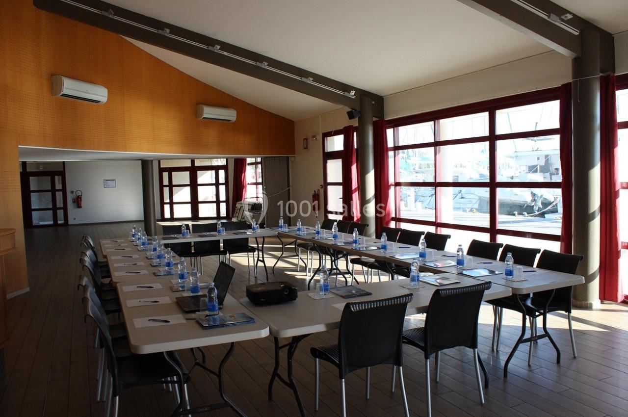 Salle de réunion lumineuse avec des tables disposées en U, équipée de chaises, bouteilles d'eau et matériel de présentation.