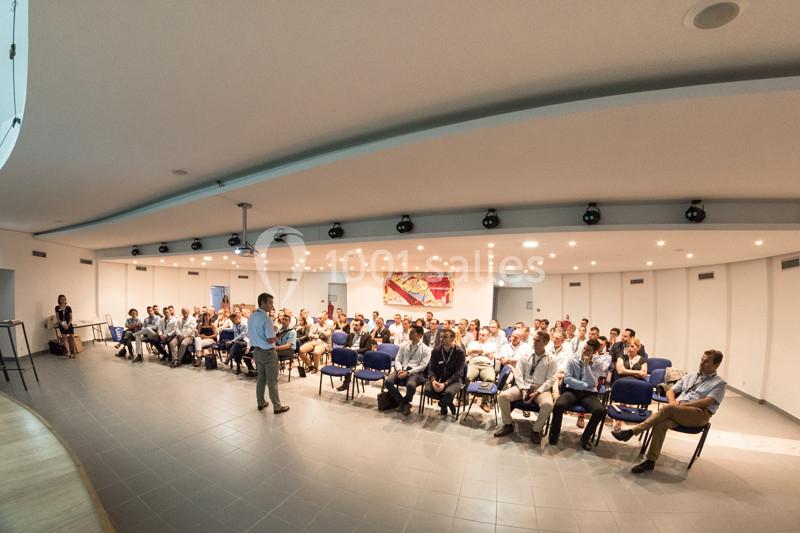 Un conférencier s'adresse à un public assis dans une salle lumineuse équipée de chaises et d'un projecteur.