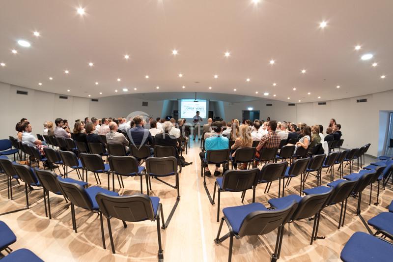 Un groupe de personnes assises dans une salle de conférence éclairée, écoutant un intervenant devant un écran.