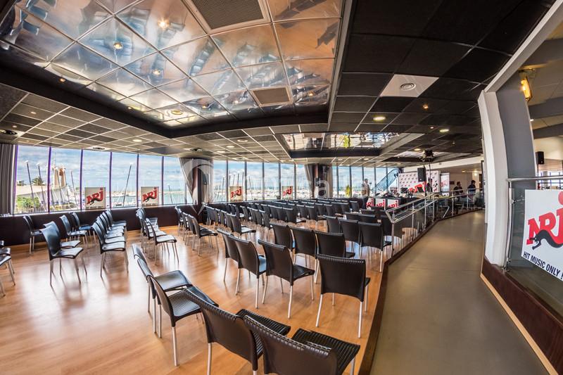 Salle de conférence lumineuse avec des rangées de chaises noires, grandes baies vitrées et plafond réfléchissant.