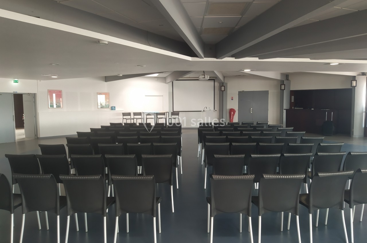 Salle de conférence vide avec des rangées de chaises noires alignées face à un écran de projection.