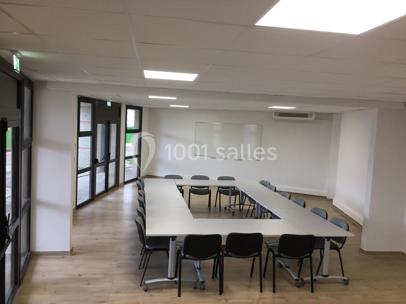 Salle de réunion lumineuse avec tables disposées en U, chaises noires, tableau blanc et sol en parquet clair.