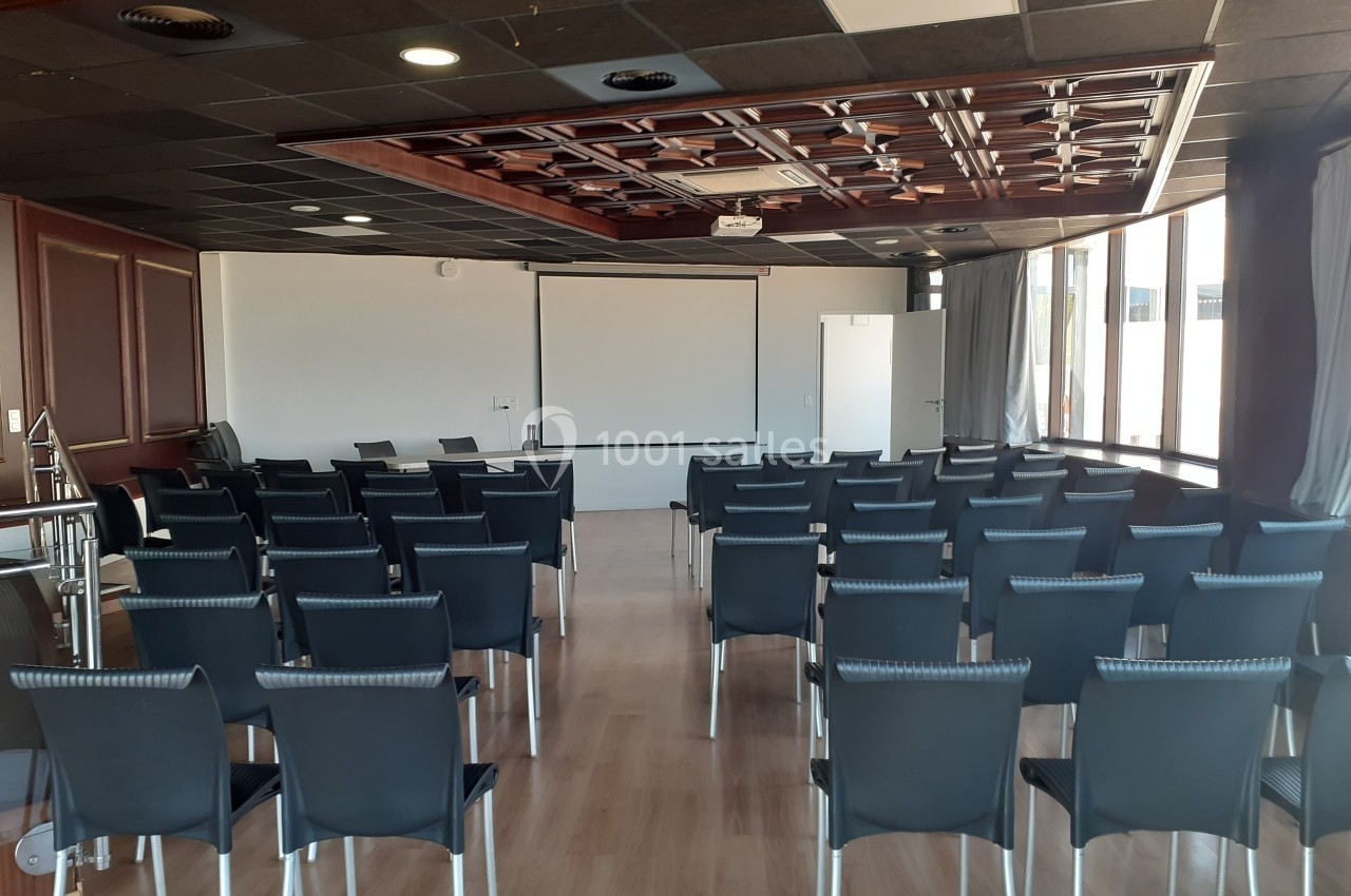 Salle de conférence vide avec des rangées de chaises noires, un écran de projection et un plafond décoratif en bois.