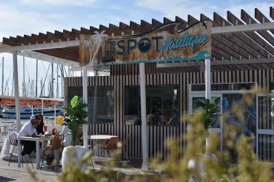 Terrasse d'un restaurant en bois avec vue sur un port de plaisance, tables occupées et végétation en premier plan.
