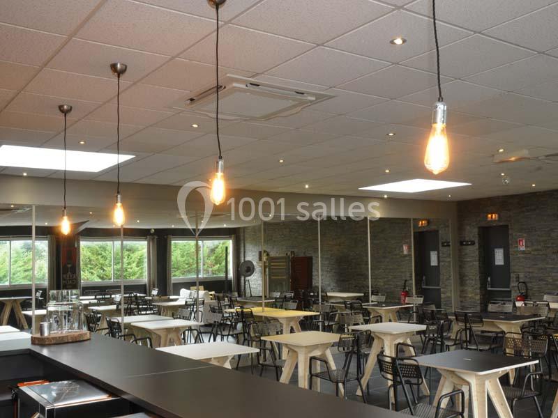 Salle de restaurant moderne avec tables, chaises noires et éclairage suspendu, murs en pierre et grandes fenêtres.