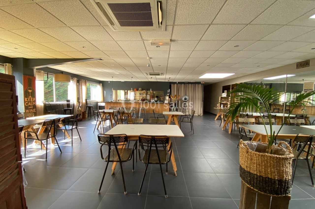 Salle lumineuse avec tables et chaises, décorée de plantes et d'éléments en bois, vue sur un comptoir en arrière-plan.