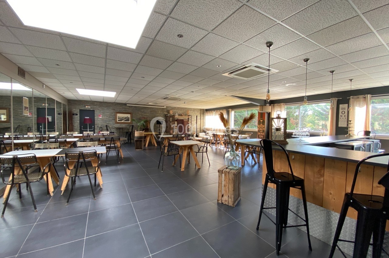 Salle lumineuse avec tables, chaises, bar en bois et grandes fenêtres donnant sur un espace extérieur verdoyant.