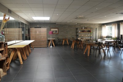 Salle lumineuse avec tables en bois, chaises noires, sol en carrelage sombre et murs en pierre.