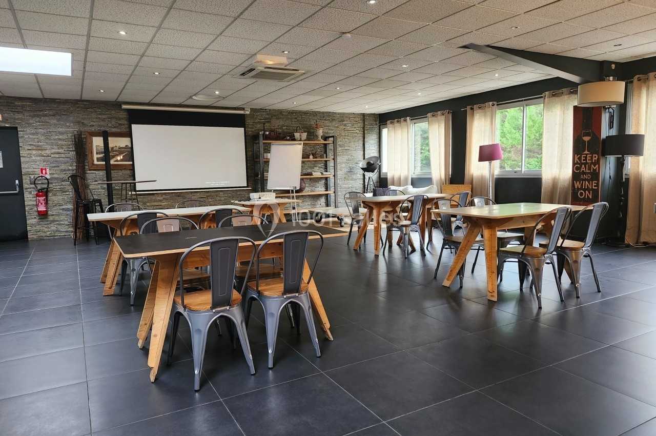 Salle lumineuse avec tables en bois, chaises métalliques, écran de projection et grandes fenêtres avec rideaux beiges.