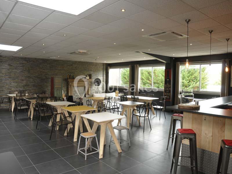 Salle lumineuse avec tables et chaises en bois, murs en pierre et grandes fenêtres donnant sur un espace extérieur verdoyant.