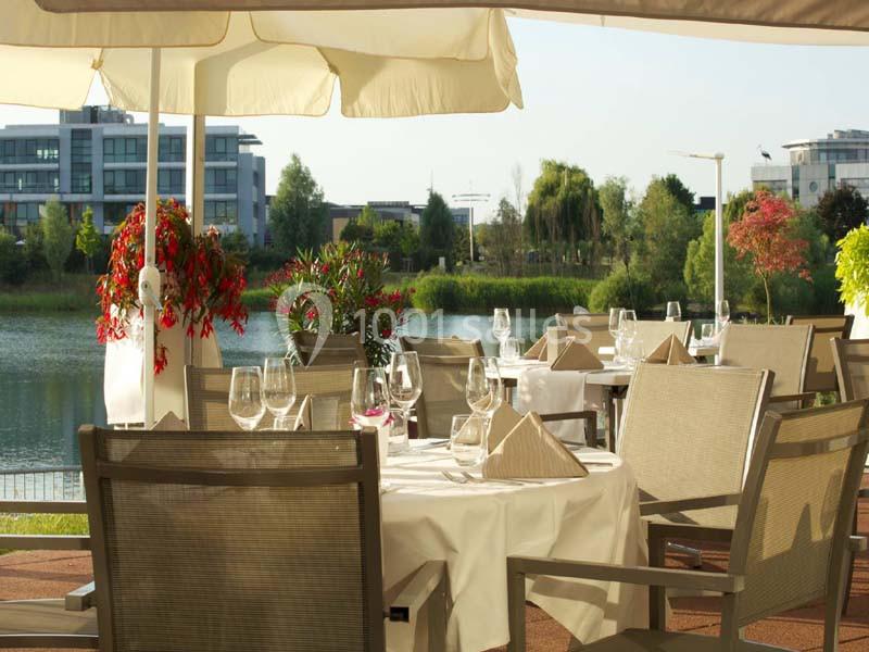 Terrasse de restaurant avec tables dressées, vue sur un lac entouré de verdure et bâtiments modernes en arrière-plan.