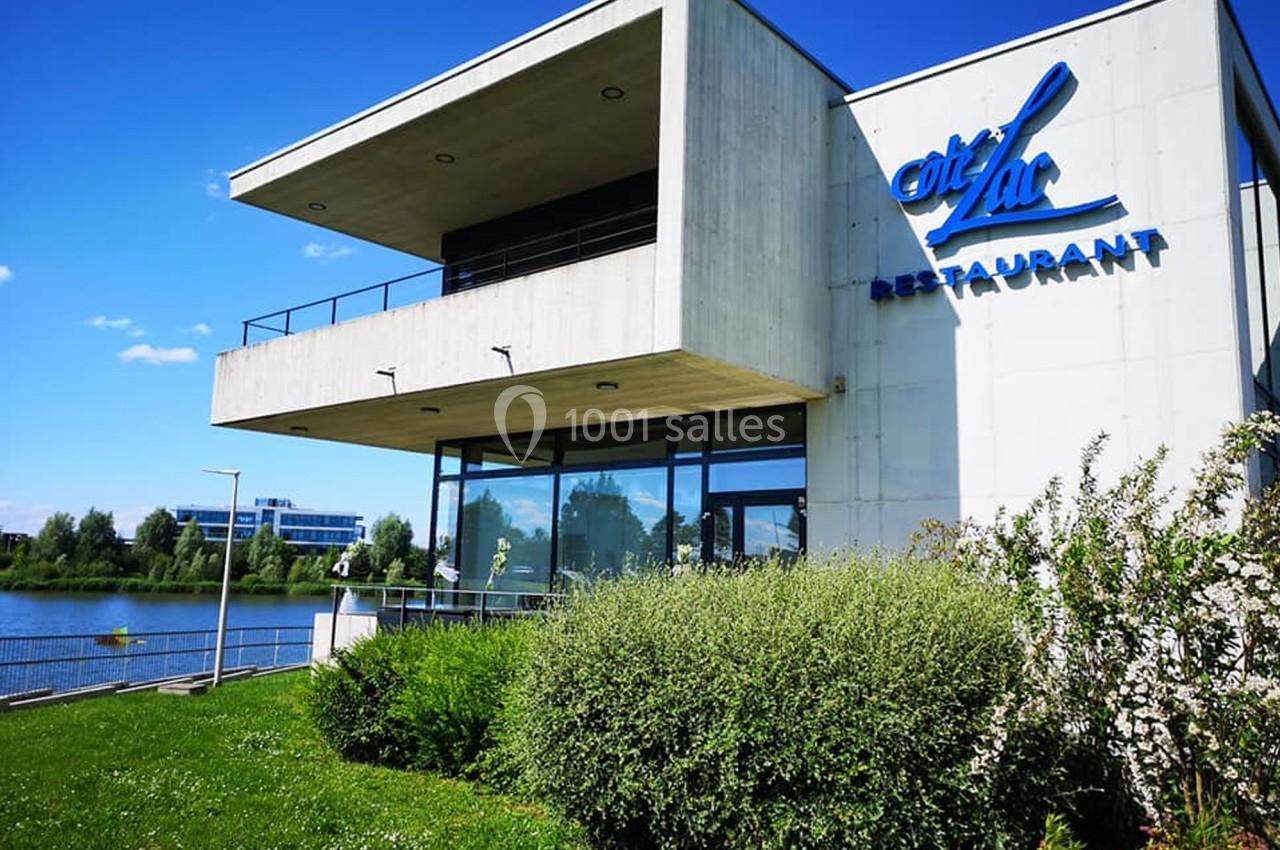 Bâtiment moderne avec terrasse, abritant un restaurant, situé au bord d’un lac par une journée ensoleillée.