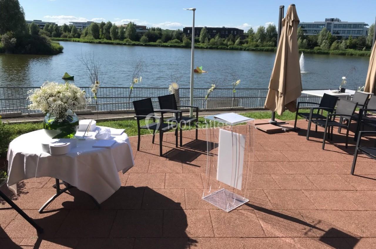 Terrasse ensoleillée avec vue sur un lac, tables dressées et parasols fermés, cadre calme et verdoyant.
