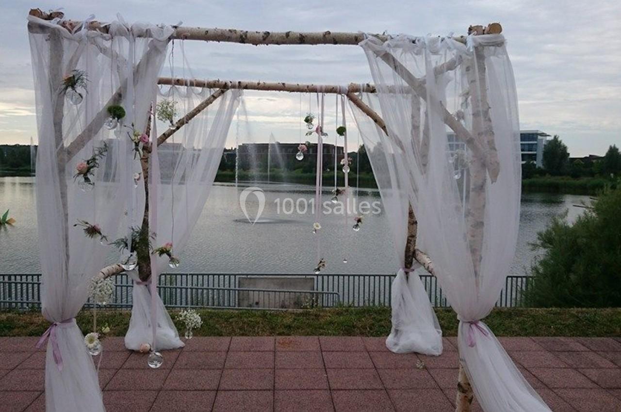 Arche de mariage en bois décorée de voilages blancs et de fleurs, installée devant un lac paisible.