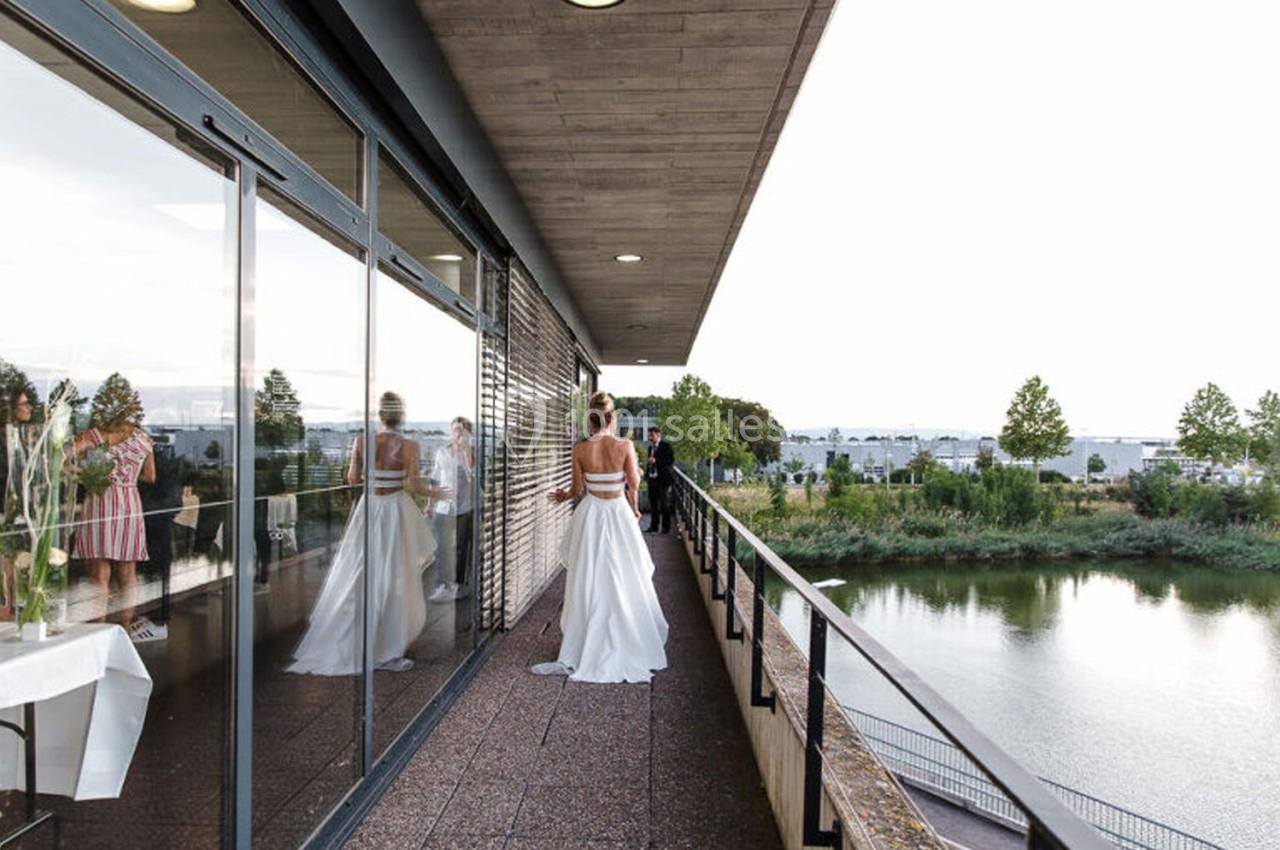 Une femme en robe blanche marche sur une terrasse longeant un bâtiment vitré, avec vue sur un plan d'eau et des arbres.