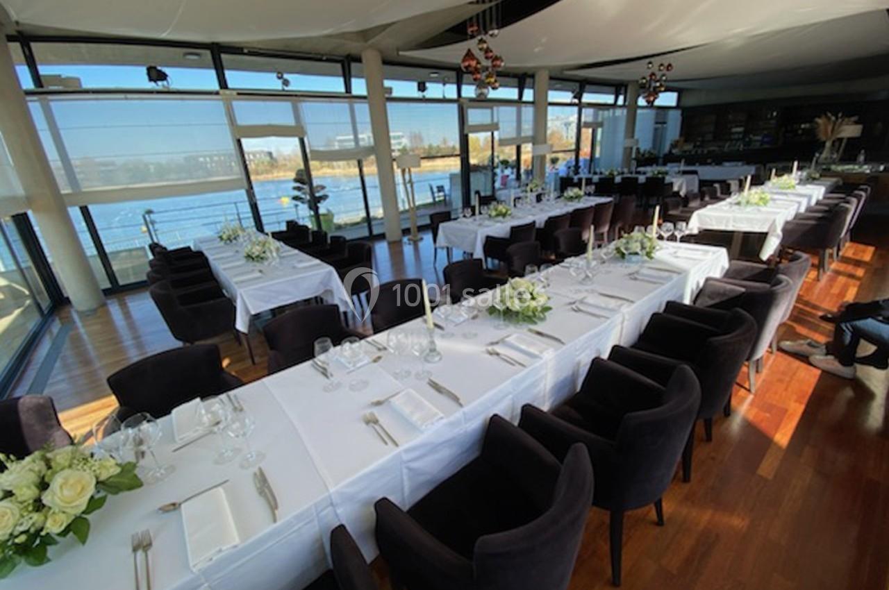Salle lumineuse avec grandes baies vitrées, tables dressées avec nappes blanches, chaises noires et décorations florales.