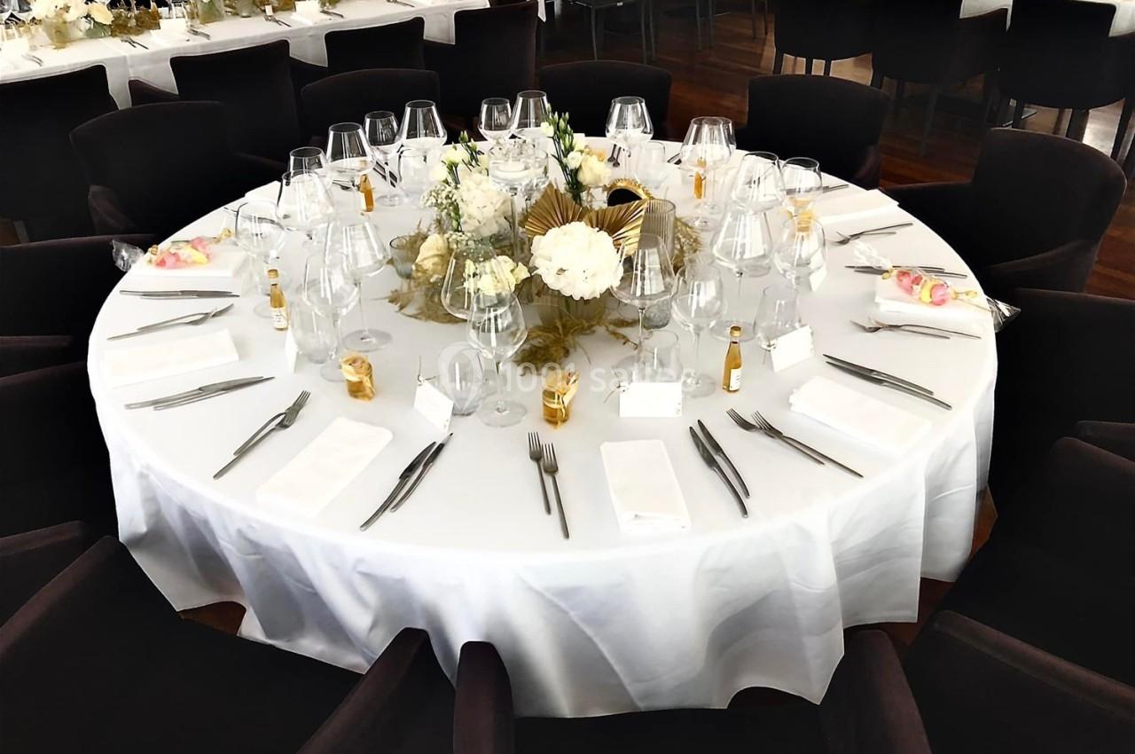 Table ronde élégamment dressée avec nappe blanche, verres, couverts, décorations florales et chaises noires.