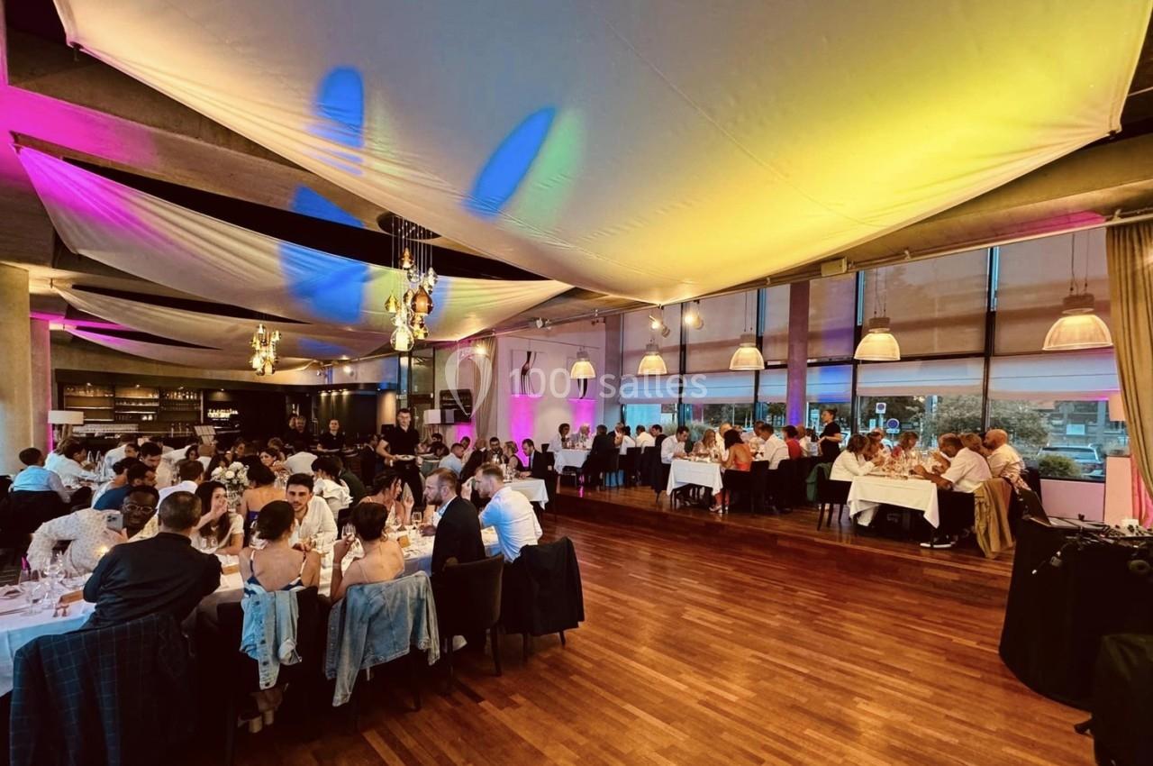 Salle de réception animée avec des invités assis à des tables rondes, éclairage coloré et ambiance festive.