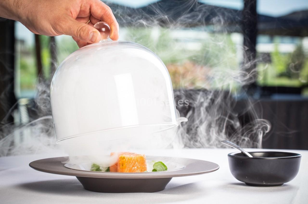 Une main soulève une cloche en verre laissant échapper de la fumée au-dessus d'un plat gastronomique sur une table.