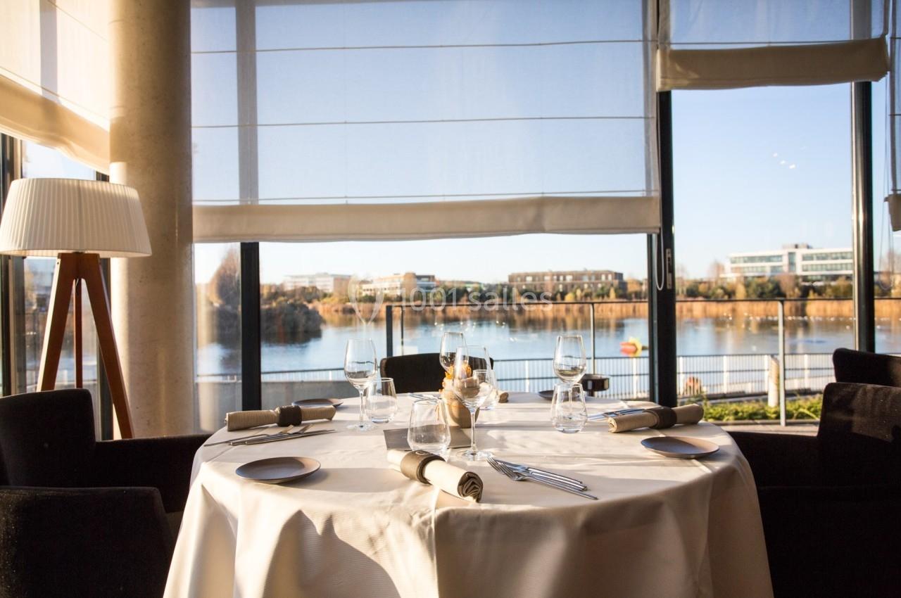 Table dressée avec élégance près d'une baie vitrée offrant une vue sur un lac et des bâtiments au loin.