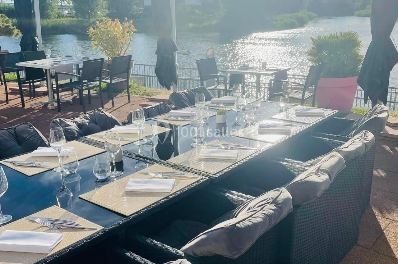 Table dressée en extérieur avec vue sur un lac, entourée de chaises en rotin et protégée par des parasols.