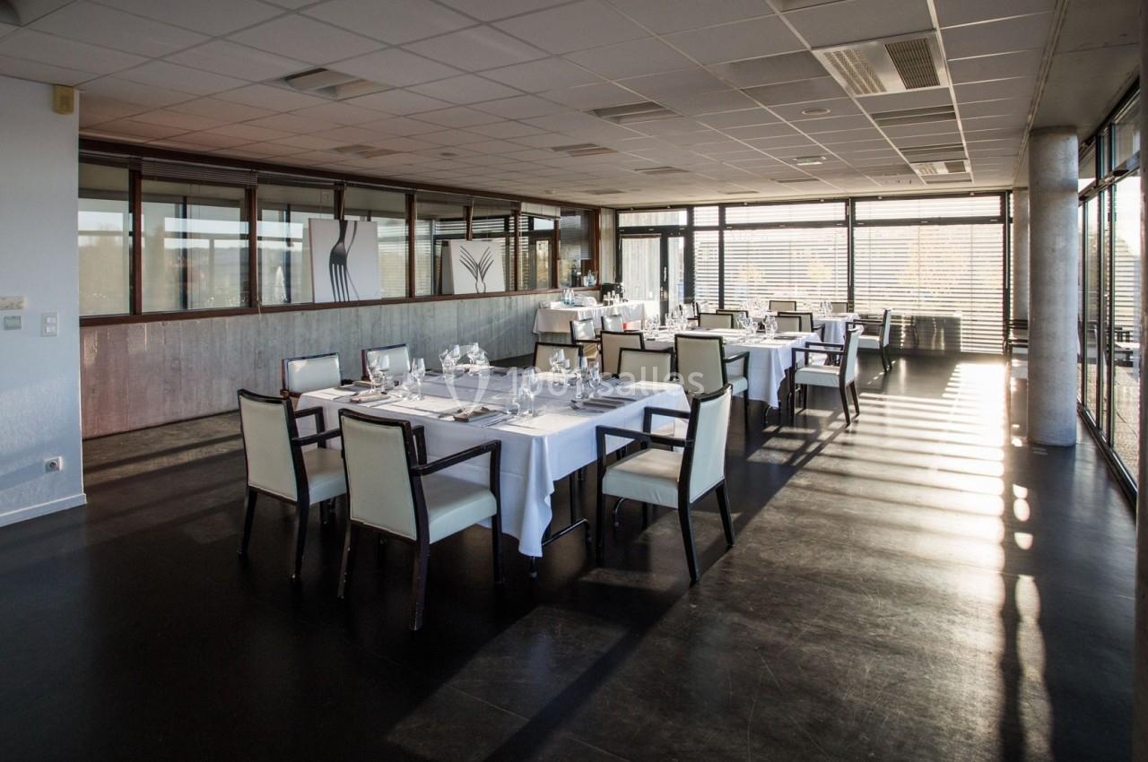 Salle de restaurant lumineuse avec tables dressées, chaises blanches et grandes baies vitrées laissant entrer la lumière…