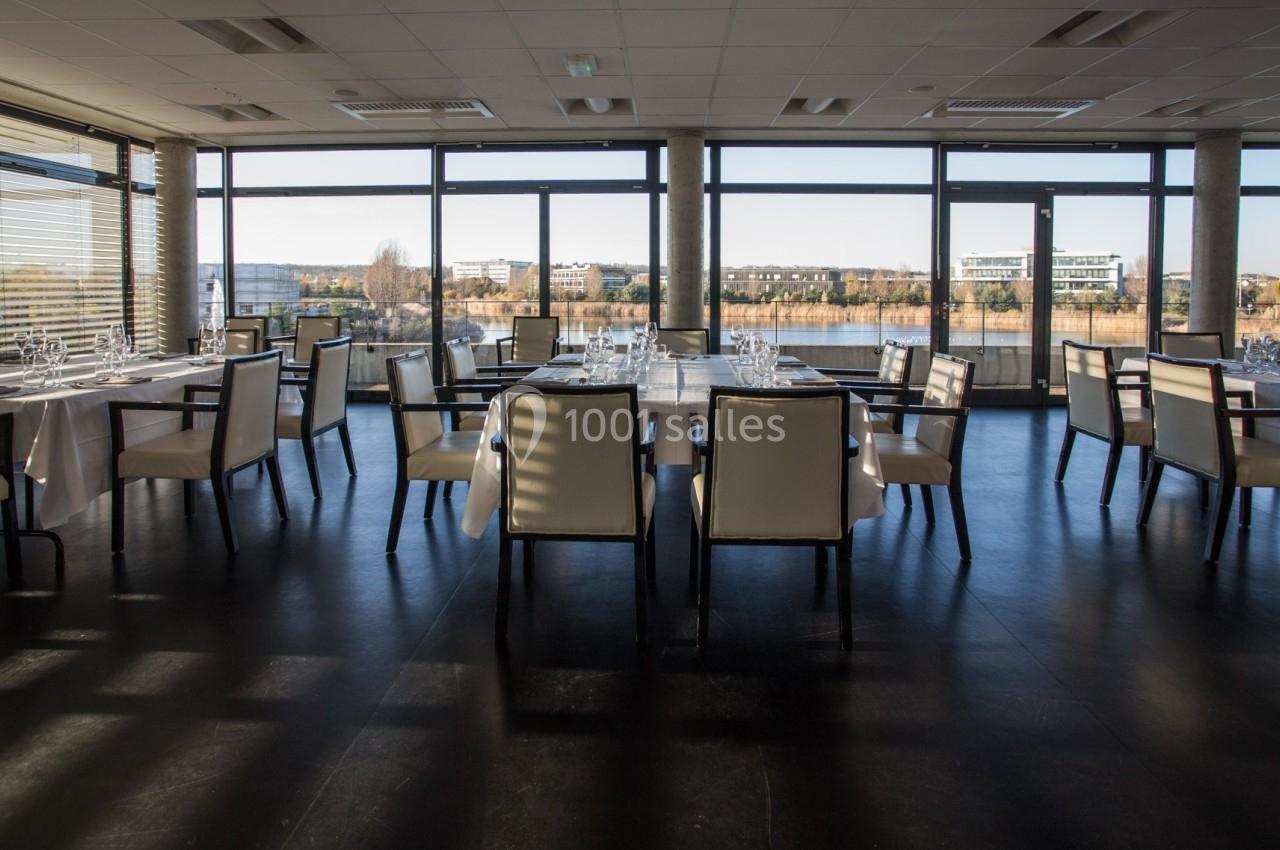 Salle de restaurant moderne avec grandes baies vitrées offrant une vue sur un paysage urbain et un cours d'eau.