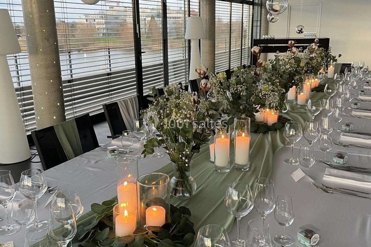 Table élégamment dressée avec nappes blanches, bougies, fleurs et décoration suspendue dans une salle lumineuse.