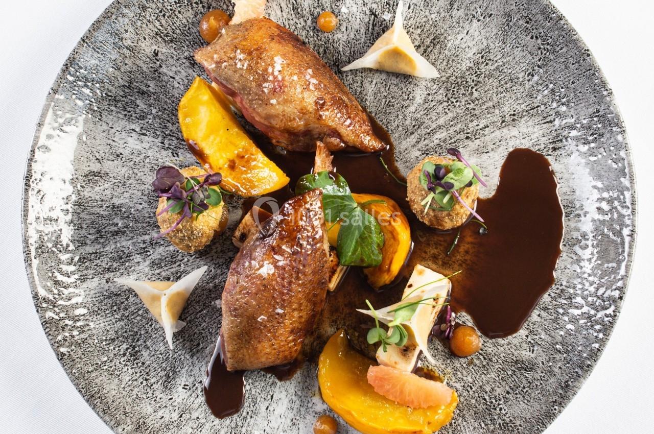 Assiette gastronomique avec viande rôtie, morceaux de fruits, sauce brune et garnitures végétales sur une assiette texturée.