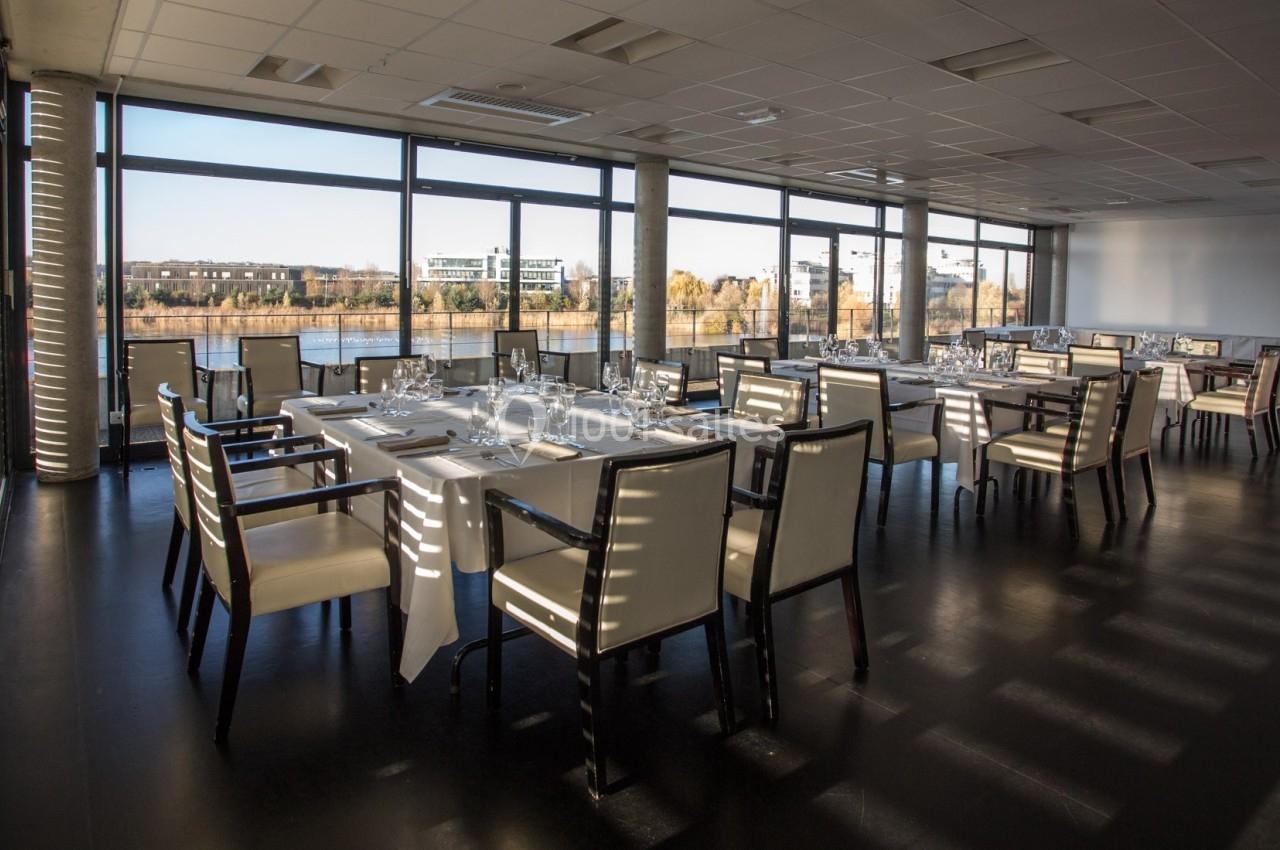 Salle de restaurant lumineuse avec grandes baies vitrées offrant une vue sur un paysage urbain et fluvial.