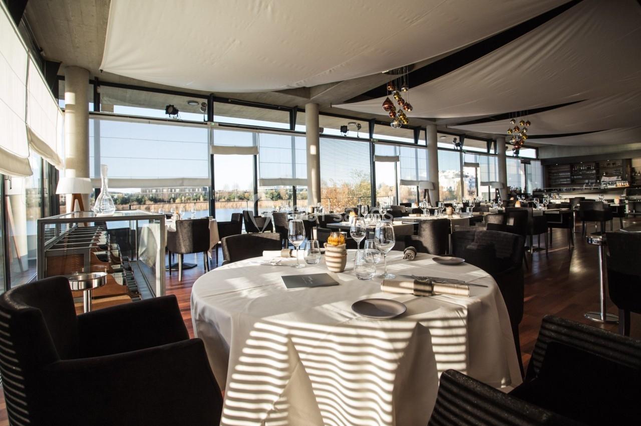 Salle de restaurant lumineuse avec grandes baies vitrées, tables dressées et vue sur l'extérieur.